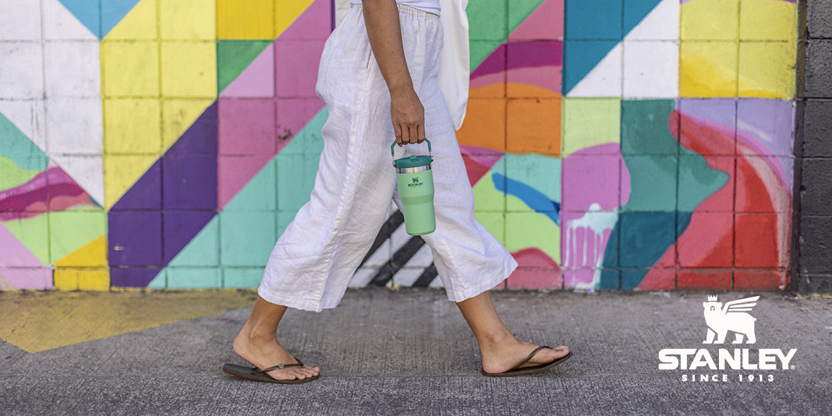 Person carrying a green Stanley water bottle walking past a colorful outdoor mural.