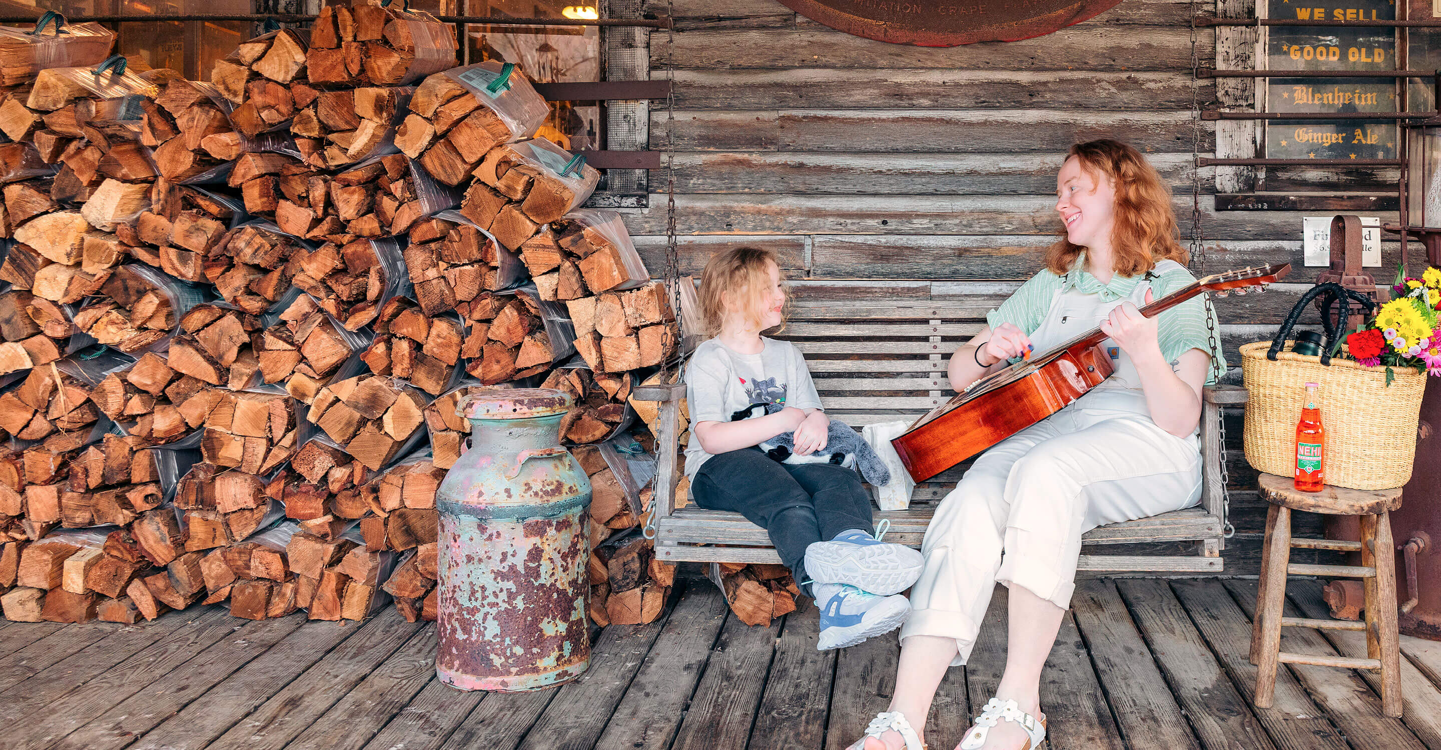 A mother and daughter sitting on a wooden swing beside stacked firewood on the back porch of the Original Mast General Store in Valle Crucis, North Carolina, smiling and looking at each other as the mother plays a guitar, with a nearby stool holding a basket of spring flowers and an open glass bottle of Nehi